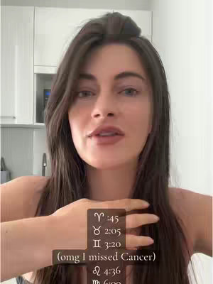 A woman with long dark hair is positioned in front of a neutral background which appears to be a kitchen. She's facing the camera with a slightly downward gaze, her hands holding a vertical label with astrological symbols and times. The overall composition is a medium shot, highlighting her face, upper body, and the overlaid text.