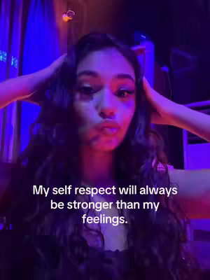 A woman with long, dark wavy hair holds her hands up to the sides of her head. She has a neutral expression with slightly pursed lips. The background is dark, with purple and blue lighting and a suggestion of some interior setting.