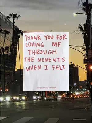 The image captures a cityscape through what appears to be a window or glass pane. In the foreground is a white paper sheet with handwritten text in red, illuminated against the backdrop of city lights, buildings, and a sunset sky. The composition is straightforward, with the text as the central focus.