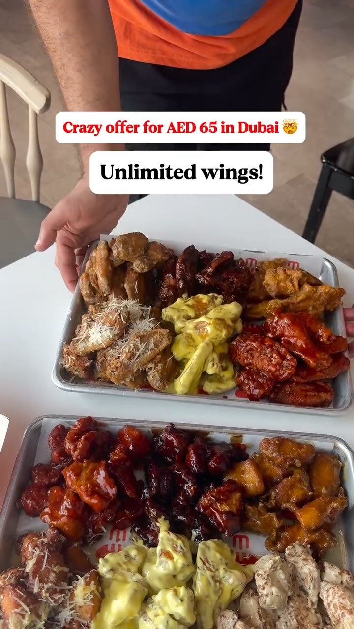 The image showcases two metal trays filled with various chicken wings, arranged to highlight different flavors. The wings are visibly coated with sauces and toppings, indicating a diverse selection. A person's hands are partially visible, holding one of the trays, suggesting a food presentation.