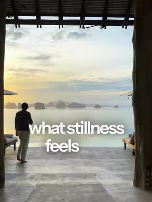 A serene image captures a moment of tranquility. A figure, seen from the back, stands looking out over a calm bay speckled with distant islands. The foreground features a covered porch with pillars, framing the view, while text overlays read "what stillness feels" across the scene.