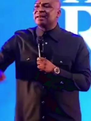 A medium shot shows a Black man in a black shirt speaking into a microphone. He has a bald head and a serious expression. The background is a blurry blue.
