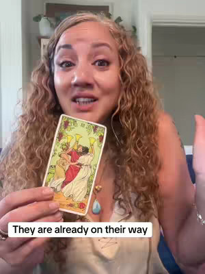 A woman with curly hair holds up a tarot card in front of her face and presents it to the viewer. Text overlayed at the bottom of the video. The background includes plants and a neutral-toned interior.