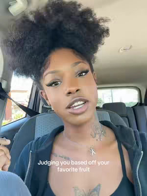 A selfie-style shot taken from inside a car, the main focus being a woman with intricately styled dark, curly hair. She has full makeup and detailed long lashes, with a tattoo on her neck. The text overlay at the bottom reads, 'Judging you based off your favorite fruit'. There are car seats and a soft-focus background, suggesting a day time shot.
