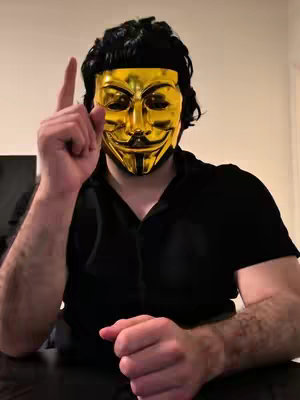 A person wearing a gold mask and black attire points upward with their index finger.