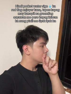 A young man in a black shirt is seen in profile, looking towards the right, with his index finger touching his lips in a shushing gesture. The background is minimal, featuring a wall on the left and what appears to be a window with a curtain on the upper right.