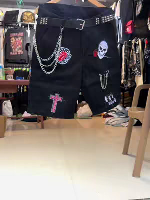 A pair of black shorts hangs on a wooden clothes hanger, prominently displayed against a backdrop of a retail environment. The shorts are heavily embellished with patches and hardware in a punk or rock-and-roll aesthetic. Notable decorations include a red cross, a Rolling Stones tongue logo, a skull with a rose, and a chain. The scene implies a clothing store or a similar retail setting, with the shorts as the primary focal point.
