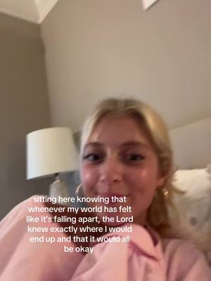 A selfie-style shot featuring a young woman with blonde hair, soft lighting, and a background suggesting a home interior. The woman is looking towards the camera with an expression of hopeful positivity. The primary colors are soft, with natural skin tones and a hint of pink in the clothing, complementing the beige-toned wall and the soft light emanating from the lamp visible in the backdrop.
