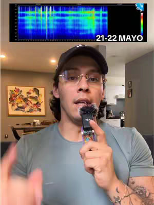 A man with glasses and a black cap holds a microphone, speaking into the camera. Above him is a colorful spectrum graph labeled with the dates "21-22 MAYO". The background includes parts of a room with a painting on the wall.