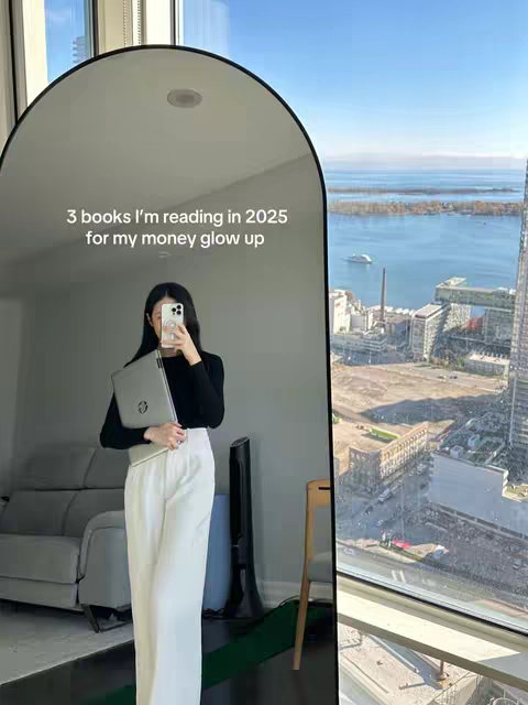 A woman stands in front of a large, arched mirror taking a photo with her phone. She is dressed in a black sweater and white pants. The mirror reflects a room with a gray couch, a view of a cityscape through a window, and text overlayed on the mirror. The text reads "3 books I'm reading in 2025 for my money glow up".