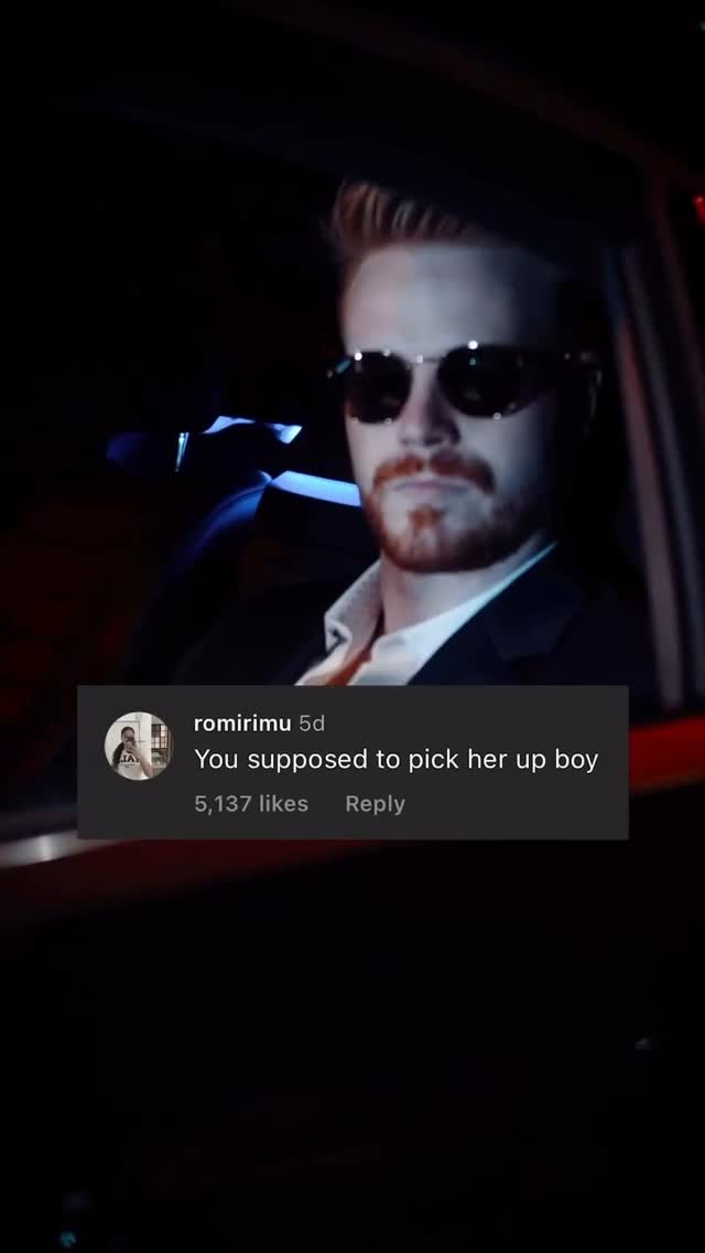 A man in sunglasses is seen from the chest up, seated in a dimly lit car. The image has a cinematic feel from some light leak and the dark setting. A social media comment is overlaid on image. The comment, displayed within a gray box, reads: "You supposed to pick her up boy."