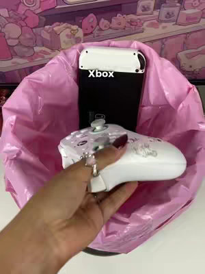 A pink trash bag sits on a white surface, containing a black device labeled "Xbox" and a white Nintendo Switch controller. A hand holding a white controller with a silver ring is in the foreground.