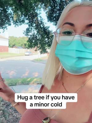 A woman wearing glasses and a turquoise face mask stands outside, appearing to be in a yard or park.  A backdrop of trees, a street, and buildings is visible. The overlaying text reads "Hug a tree if you have a minor cold".