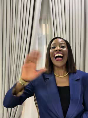 A woman with shoulder-length dark hair is waving and smiling at the camera. She's wearing a blue blazer, a black top, and a gold necklace and bracelets. The background features vertical white curtains. The overall composition has a warm and inviting feel.