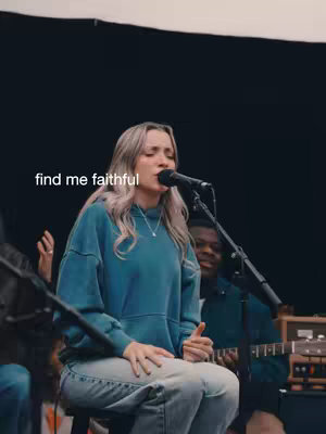 A woman with long, highlighted hair wearing a teal hoodie and jeans is singing into a microphone, sitting on a stool. The words "find me faithful" are overlaid on the image. Behind her, in a blurred background, is a band setup.