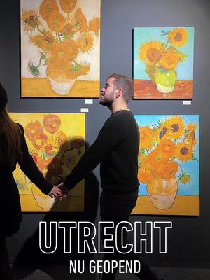 A person in a dark sweater and jeans, holding the hand of a person off-screen, looks up at four Van Gogh paintings of sunflowers hanging on a dark wall. The paintings are in the top and upper-left of the frame.  A large white text block at the bottom says 'UTRECHT' then 'NU GEOPEND' below it.