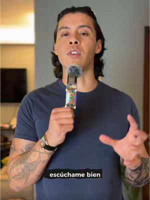 A man with dark hair holds a microphone close to his mouth, looking towards the camera with an open gaze, and his arms are slightly spread. He wears a dark blue t-shirt, and a tattoo is visible on his left arm. The background is blurred, featuring parts of a room with neutral tones.