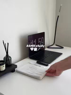 A workspace is shown with a white desk, a laptop on it opened and showing the time, a notepad and pens, and a black candle holder, and a hand to grab the notepad. In the background, out of focus, is a white wall.