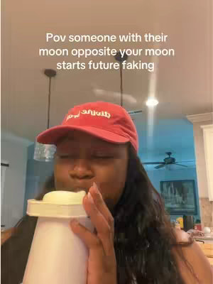 A person wearing a red baseball cap is taking a sip from a white cup in a domestic setting. The angle is low, showing the ceiling and overhead lights. The person appears to be black with long, dark hair, and is holding the cup with both hands. The background is blurred, with kitchen elements visible.