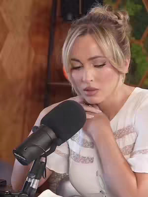 A young woman with blonde hair in a bun sits in front of a microphone, with her eyes closed. She is wearing a white top, and her hands are near her face.