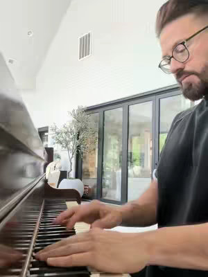 A man wearing glasses plays the piano, with his hands visible on the keys and a portion of the piano visible. The background includes a bright room with windows and a tree.