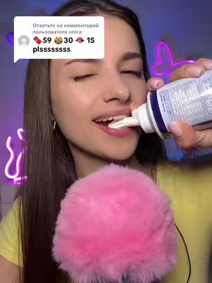 A young woman with long, dark hair is holding a can of cream and squeezing it into her mouth. She is smiling and wearing braces. A pink fluffy microphone is positioned in front of her, and there is a glowing neon butterfly outline behind her. A text bubble with comments is present at the top left corner.
