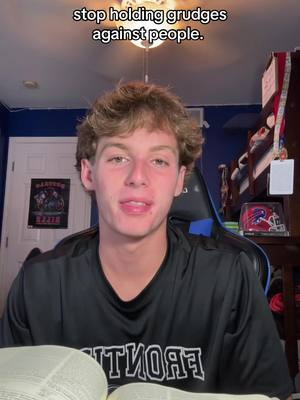 A young man with light brown hair is centered in the frame, looking directly at the camera with his mouth slightly open. Above his head, text reads 'stop holding grudges against people.' He is wearing a black shirt, and there is an open book visible in the bottom of the frame. The background features a dark blue wall, a ceiling light, and some items on a shelf, adding depth.