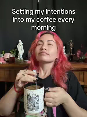 A woman with bright pink hair holds a mug of coffee, stirring it with a spoon. The image has a warm tone with a dark background. Text overlayed reads: 'Setting my intentions into my coffee every morning.'