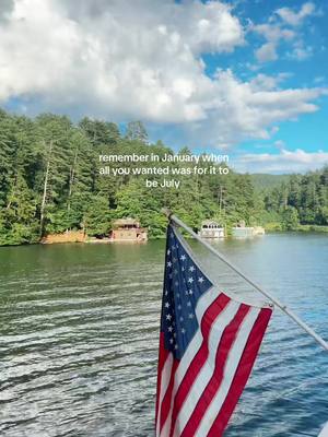 A scenic view from a boat on a lake. The image includes trees along the shoreline, a cloudy sky overhead, and an American flag waving in the foreground. Text overlay reads, 'remember in January when all you wanted was for it to be July.'