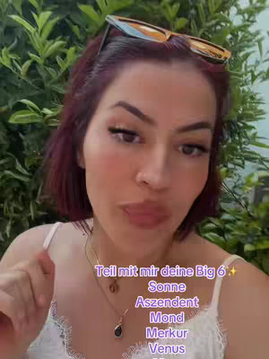 A selfie-style shot featuring a woman with dark pink hair and sunglasses perched atop her head, set against a backdrop of green foliage. The woman is looking towards the camera, possibly talking, with text overlays about astrology topics.