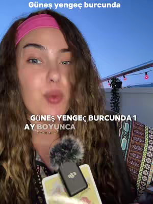 A woman with long curly brown hair and a pink headband is the central figure, facing the camera. Text overlays at the top and bottom say 'Güneş yengeç burcunda' and explain the content related to astrology. A microphone with a fluffy cover is in front of her. The background shows a blue sky. She appears to be speaking, with her lips pursed.