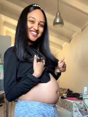 The cover image features a smiling pregnant woman with long dark hair, wearing a black long-sleeve shirt and grey shorts. She is indoors, possibly in a kitchen, with a light-colored background. She is holding her hair with one hand and giving a thumbs up with the other, while her pregnant belly is visible.