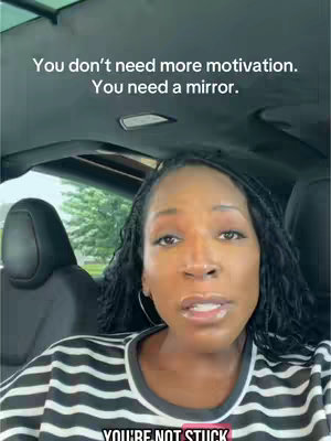 A woman is seated inside a car, looking towards the camera and speaking. Above her head is a text overlay. Her hair is styled with small braids. She wears a striped black and white shirt.