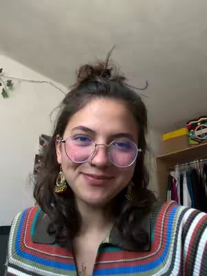 A woman with curly brown hair and round, pink-tinted eyeglasses smiles at the camera. She wears a colorful striped shirt and gold earrings. The background includes glimpses of a room with a hint of a closet.