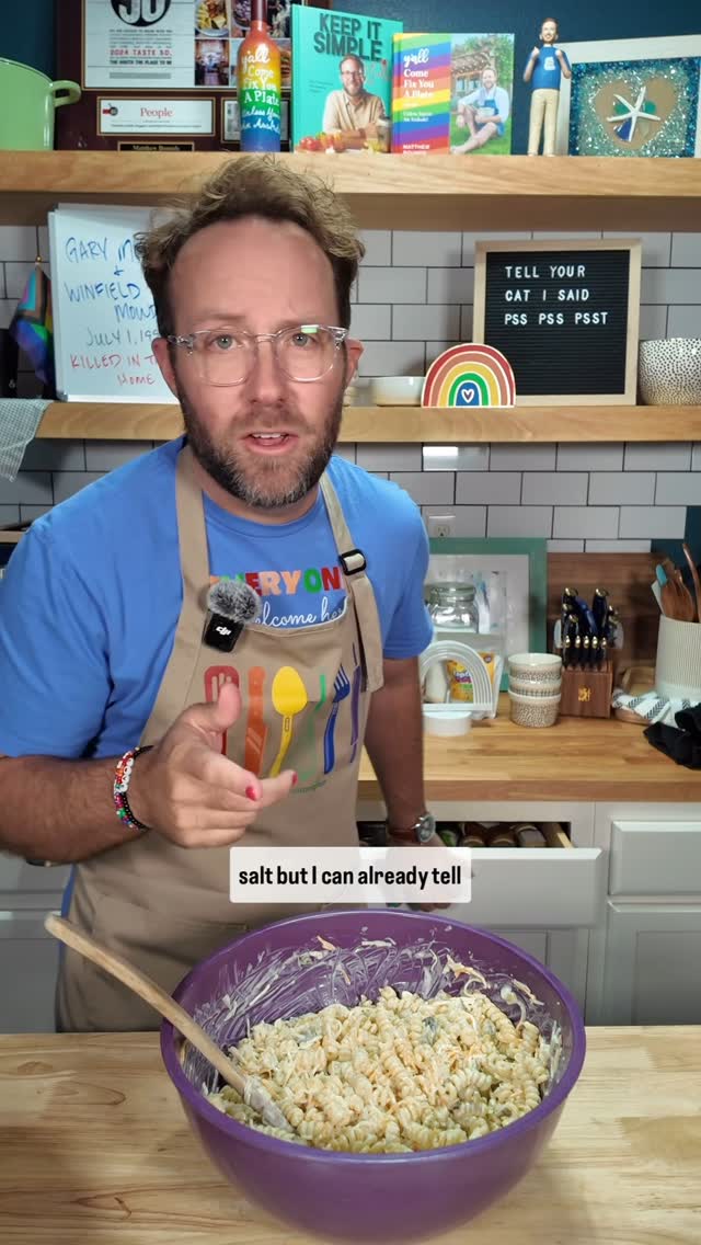A man wearing glasses, a blue t-shirt, and an apron stands in a kitchen, pointing towards a bowl of pasta salad. The background includes shelves with various items like books and decorative pieces.