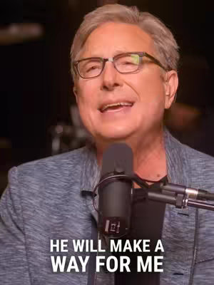 A medium shot of a man in glasses speaking into a microphone. He is the focal point, with a black microphone in front of him. The background is dark, and the text overlay reads, 'HE WILL MAKE A WAY FOR ME.' The man is wearing a blazer over a black shirt.