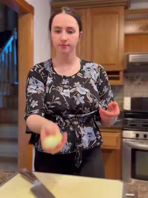 A woman in a kitchen setting, holding an onion. She wears a floral top against a backdrop of wooden cabinets.