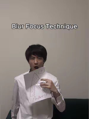 A young man with dark hair and a white collared shirt holds up a piece of paper with handwritten notes and an illustration while looking directly at the camera with an open mouth. The image is set against a plain, light-colored wall. Text overlay in the top of the image.