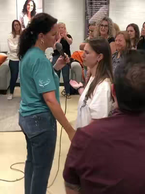 A woman in a blue shirt and jeans is speaking into a microphone, facing a younger woman who is being ministered to. Other people are in the background.