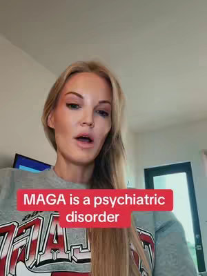 A woman with blonde hair is speaking with an open mouth. Overlay text states "MAGA is a psychiatric disorder" in white font on a red bar. She is wearing a grey sweatshirt partially visible with text on it. Behind her, a wall, a small TV, and a dark closed door are visible.