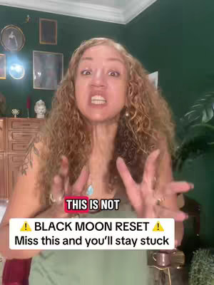 A medium shot featuring a woman with long, curly, light-brown hair. She is in a room with dark green walls and a decorative frame in the background. The woman has a direct gaze and an intense facial expression, with her hands up in a gesture. There is an overlay at the bottom with red and yellow text that emphasizes a black moon reset.
