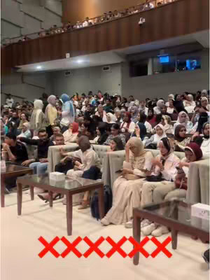 An image taken inside a large auditorium. The viewpoint is from the rear. The auditorium is filled with chairs and tables. It is occupied by a varied and large audience, mostly dressed in modest clothing, with many wearing head coverings. Red 'X' marks are overlaid on the bottom of the image.