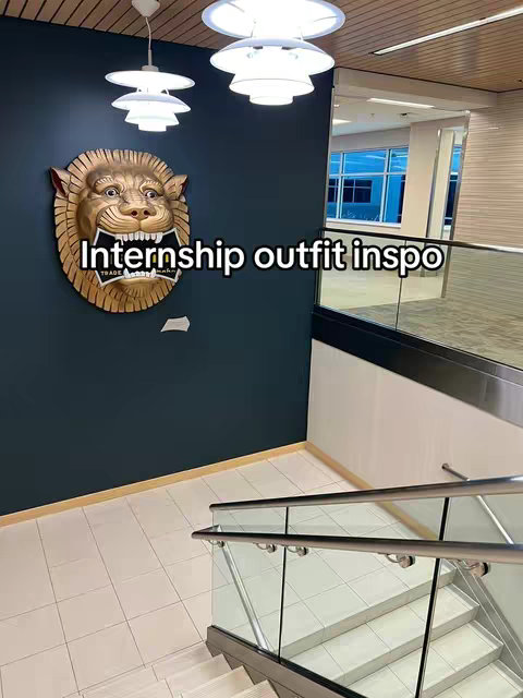 The image captures an interior scene with an office-like environment. The foreground features a staircase with glass railings, leading upwards, bathed in light. The background comprises a dark teal wall, a golden lion head sculpture, and a glimpse of a hallway.