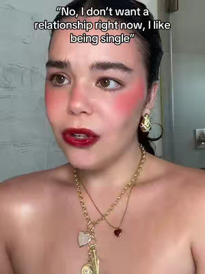 A woman with dark hair and bold makeup (bright red blush and lipstick) is looking directly at the camera. She's wearing gold jewelry, including earrings and a necklace with heart pendants. The background appears to be a bathroom setting.