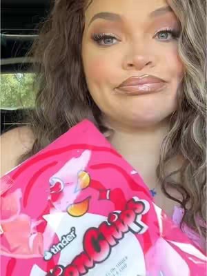 A woman with long, wavy, light brown hair holds a pink bag of "Tinder Bon Bons" that is visible over her midsection. She has full makeup on and is looking at the camera with pink highlights on her cheeks. The background is blurred, suggesting she is in a car or other enclosed space.