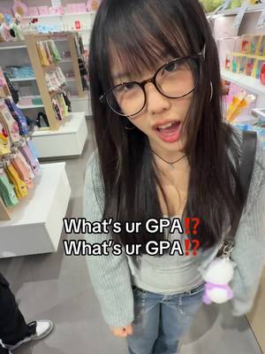 A young woman with glasses looking directly at the camera with an open mouth, in what appears to be a store setting. Text overlays read "What's ur GPA?!"