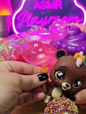 A person's hands are holding a brown bear-shaped toy and a yellow stick. The background includes other toys, various bubbles and a neon sign.