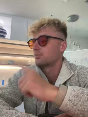 A man with blond hair and sunglasses is looking slightly to the side, with his arm in the foreground. The background has a minimalist modern design.