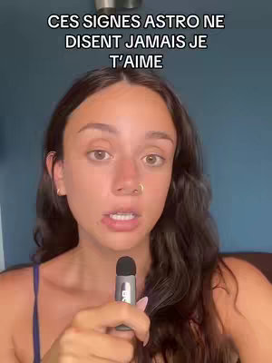 A woman with a nose ring and dark hair, is center frame holding a microphone. She is looking at the viewer with a subtle expression as text reading "CES SIGNES ASTRO NE DISENT JAMAIS JE T'AIME" is laid out above her head.