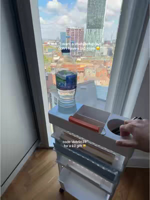 A tall, clear storage cart is positioned in front of a window with a city view. A water bottle sits on the cart along with some pens and a red item, possibly a phone charger. Text overlays are present, advertising a study setup and a Temu discount code.